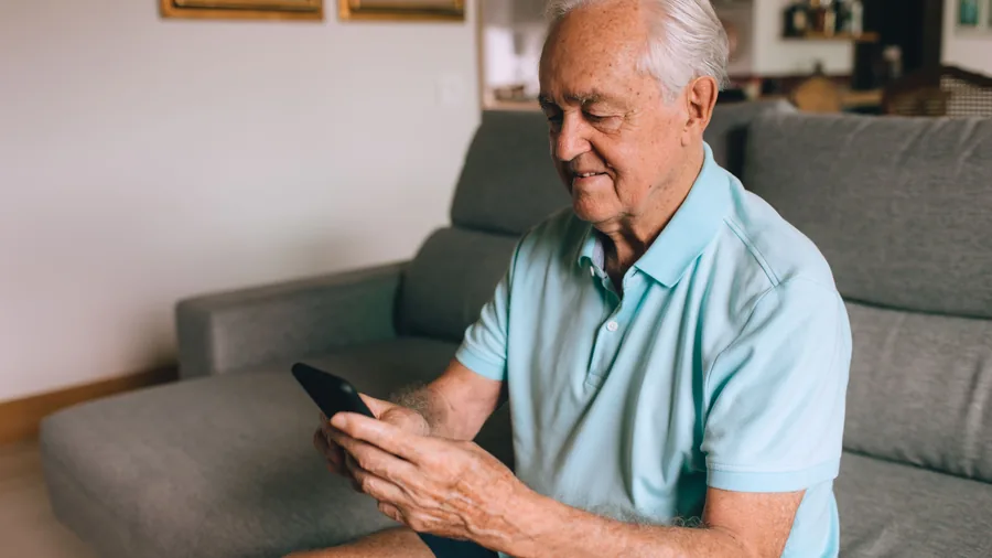 Älterer Mann sitzt auf dem Sofa und prüft mit dem Smartphone seine Unterlagen zur Pflegekasse Älterer Mann sitzt auf dem Sofa und prüft mit dem Smartphone seine Unterlagen zur Pflegekasse